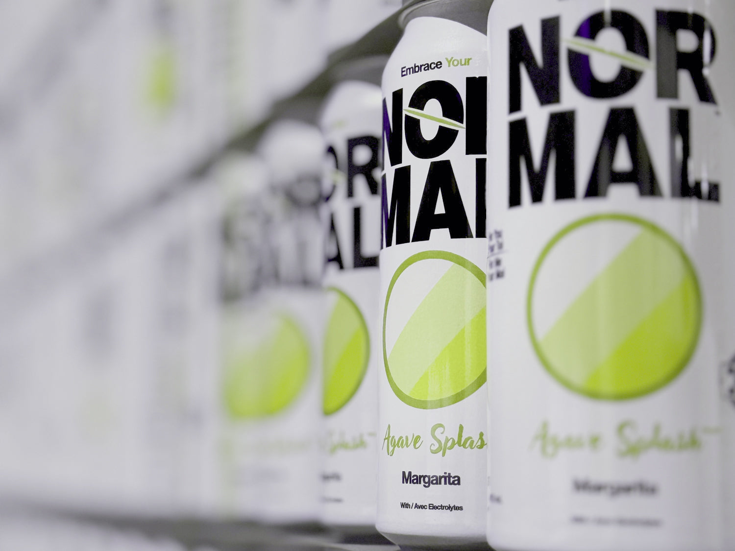 Close-up row of Normal Agave Splash Margarita cans lined up on a shelf, with the front can in sharp focus and the rest fading into a blurred background.