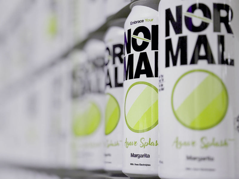 Close-up row of Normal Agave Splash Margarita cans lined up on a shelf, with the front can in sharp focus and the rest fading into a blurred background.
